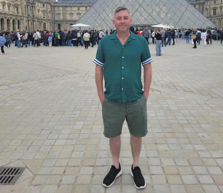 A man in a green shirt and shorts standing in front of the Louvre Pyramid in Paris.