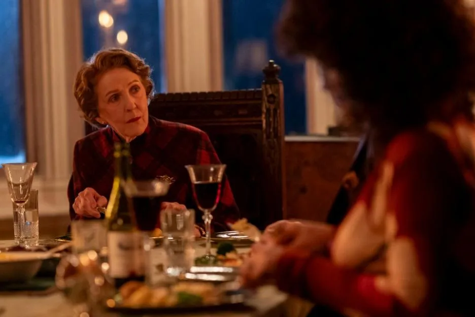 Patricia Hodge as Miriam Crace in a plaid blazer sitting at a formal dinner table with another person.