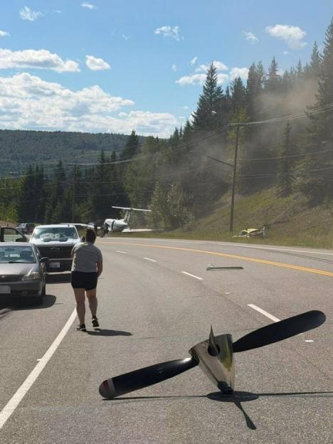 A detached plane propeller on a road near a crashed plane and other vehicles.