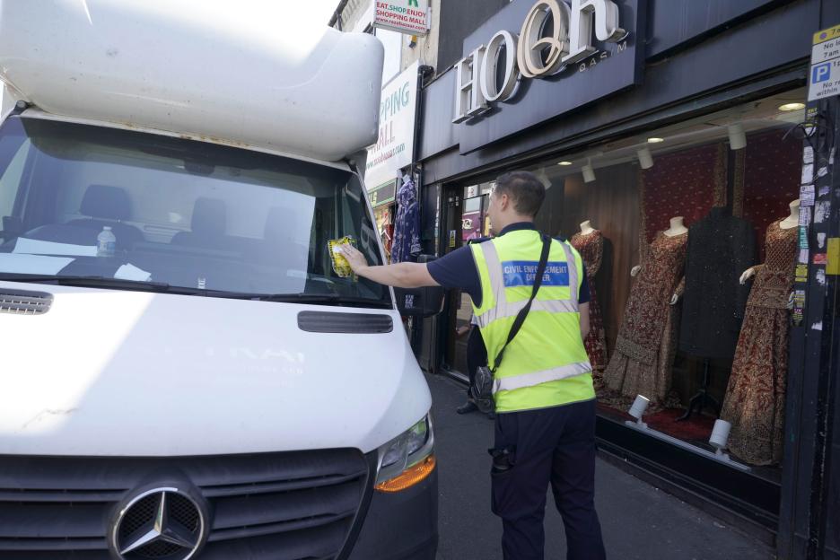 A civil enforcement officer placing a parking fine on a white van.