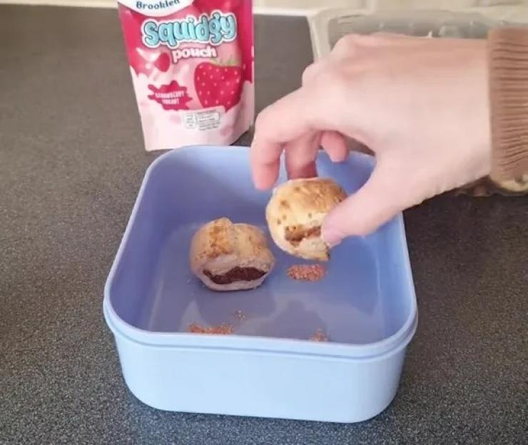 A hand places a pastry into a lunchbox with another pastry, next to a strawberry yogurt pouch.