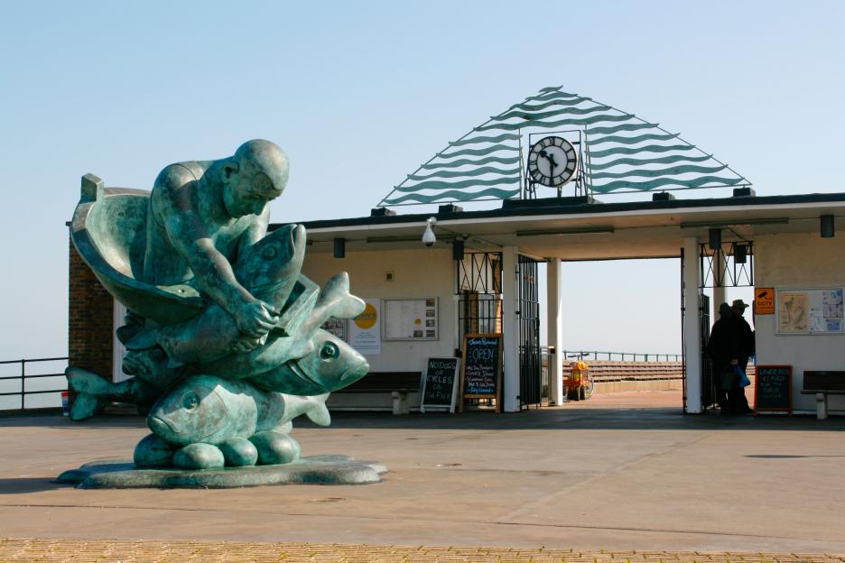 Embracing the Sea sculpture by John Buck at the entrance to Deal Pier, Kent, with a clock and a sign for a restaurant.