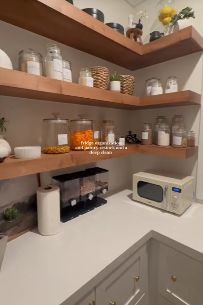 A clean and organized pantry with open wooden shelves displaying various food items in clear jars, a microwave, and a cereal dispenser.