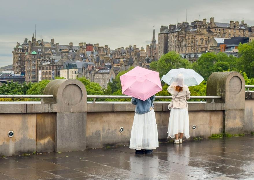 Two tourists with umbrellas overlooking the old town architecture of Edinburgh on a wet summer day.