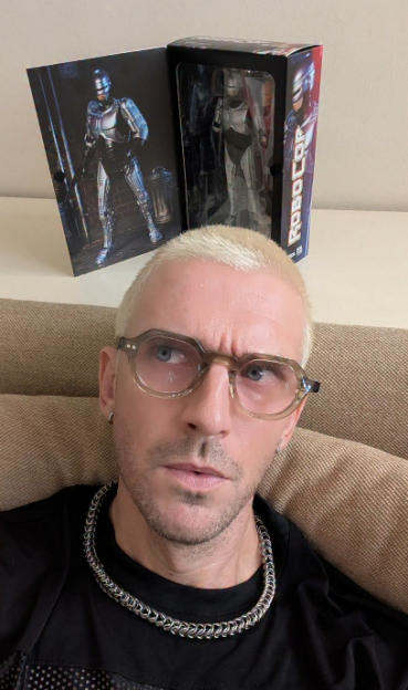 A man with blonde hair and glasses poses with a Robocop figure and a Robocop image.
