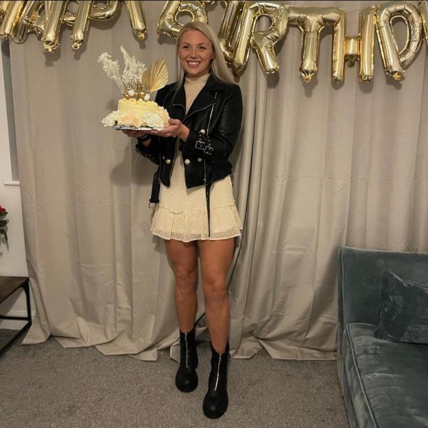 Pamela Pritchard holding a cake, wearing a black leather jacket, beige dress, and black boots, with "HAPPY BIRTHDAY" balloons in the background.