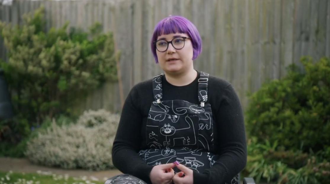 A young woman with purple hair, glasses, and a black cat-patterned overall top, sitting outdoors in a garden setting.