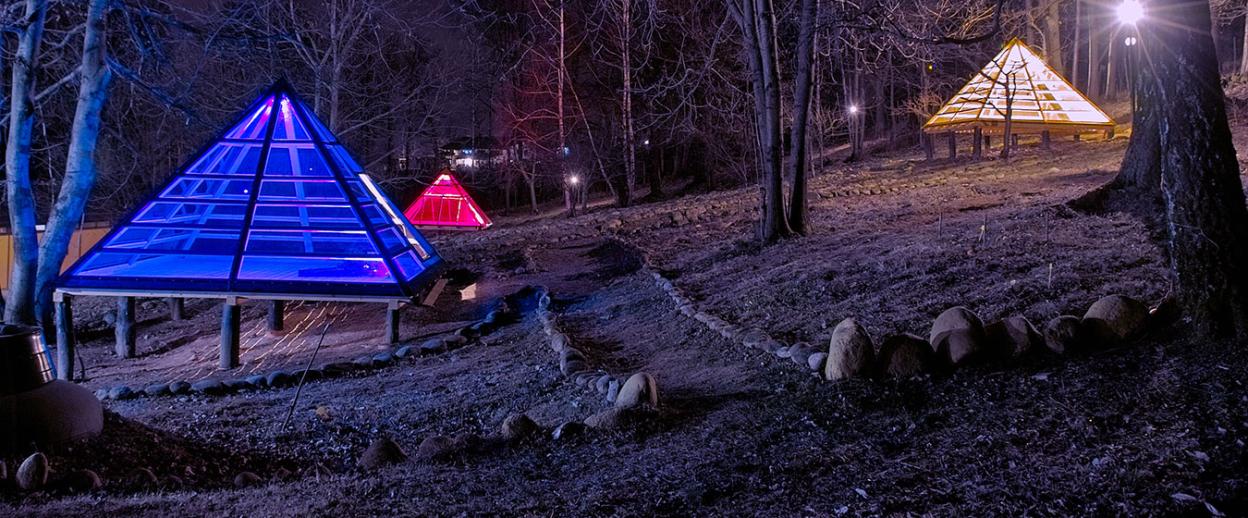 Three pyramids, one blue, one red, and one yellow, lit up at night in a wooded area with bare trees.