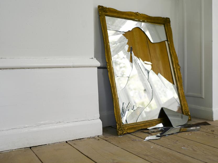 A broken mirror in a gold frame leaning against a white wall with shards on a hardwood floor.