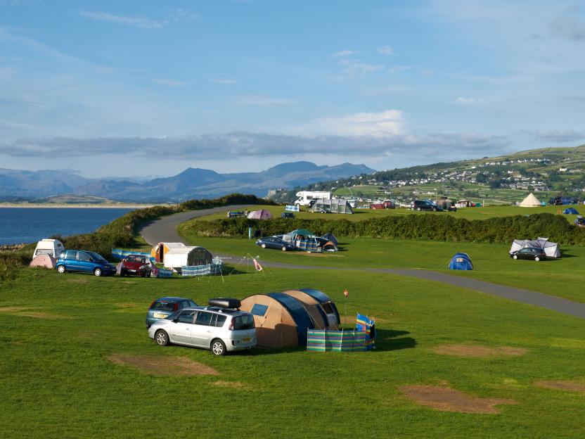 A Shell Island campsite with tents and cars on green grass, next to a road, with the sea and distant mountains in the background.