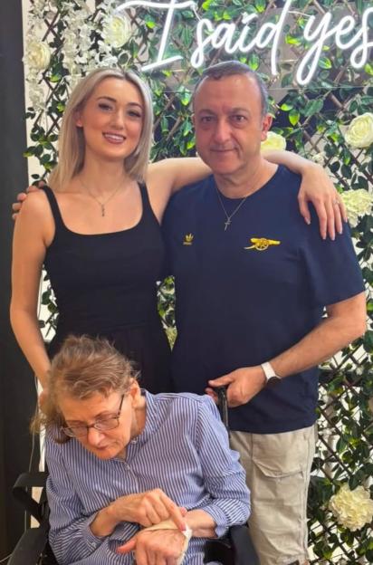 Anna, John, and Gilly posing for a photo in front of a decorative wall with white flowers and the neon sign "I said yes".