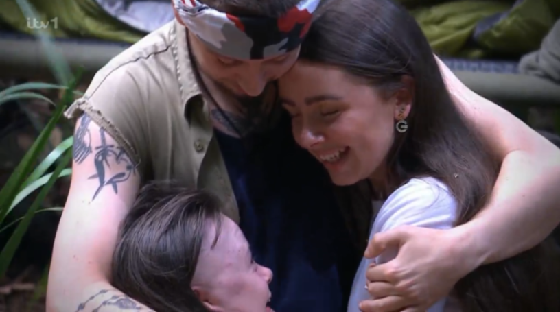 Three people embracing in a group hug, with two of them smiling and looking at each other, and the third person looking up at them.