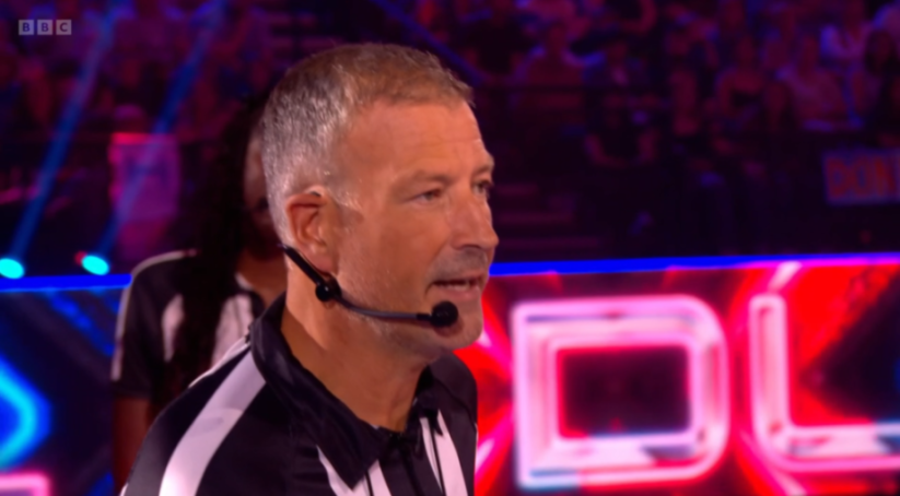 A close-up of referee Mark Clattenburg speaking into a headset, wearing a black and white striped shirt.