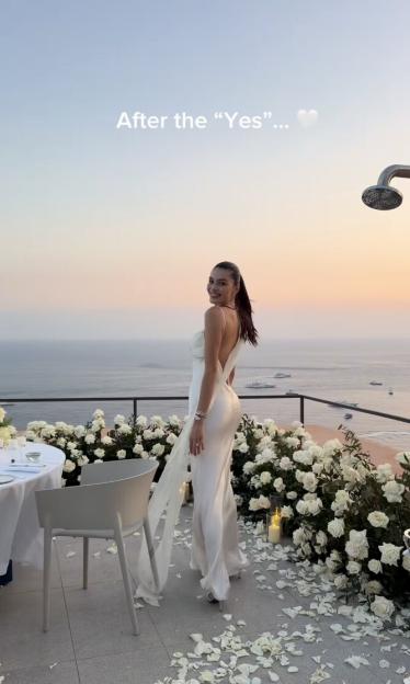 Nathalia Campos, an engaged Love Island star, smiling in a white dress on a balcony decorated with white roses overlooking the sea at sunset.