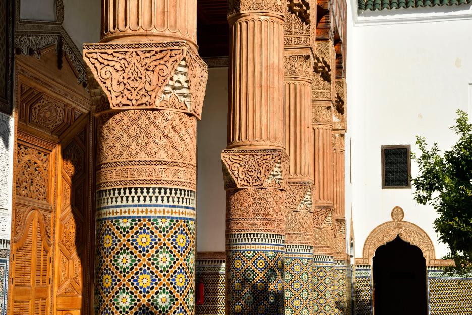 Four intricately carved columns with mosaic bases and a wooden door at the Museum of Confluences Dar El Bacha in Marrakech, Morocco.