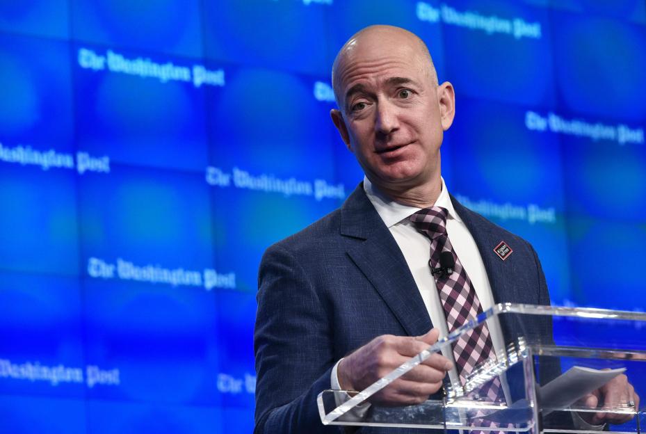 Jeff Bezos speaking at an event, with "The Washington Post" visible in the background and a "#JasonIsFree" pin on his lapel.