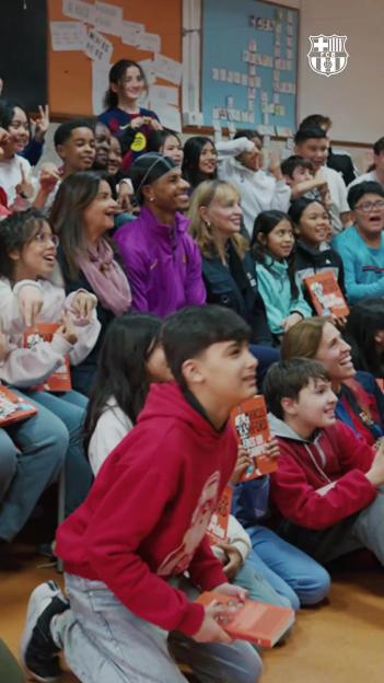 A group of children and adults sitting and kneeling, smiling and looking to the right, some holding orange books.