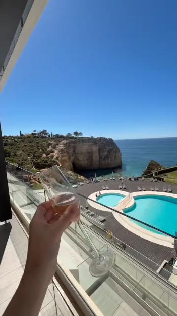 Hand holding a champagne flute on a balcony overlooking an ocean, rocky cliffs, and a resort pool area.