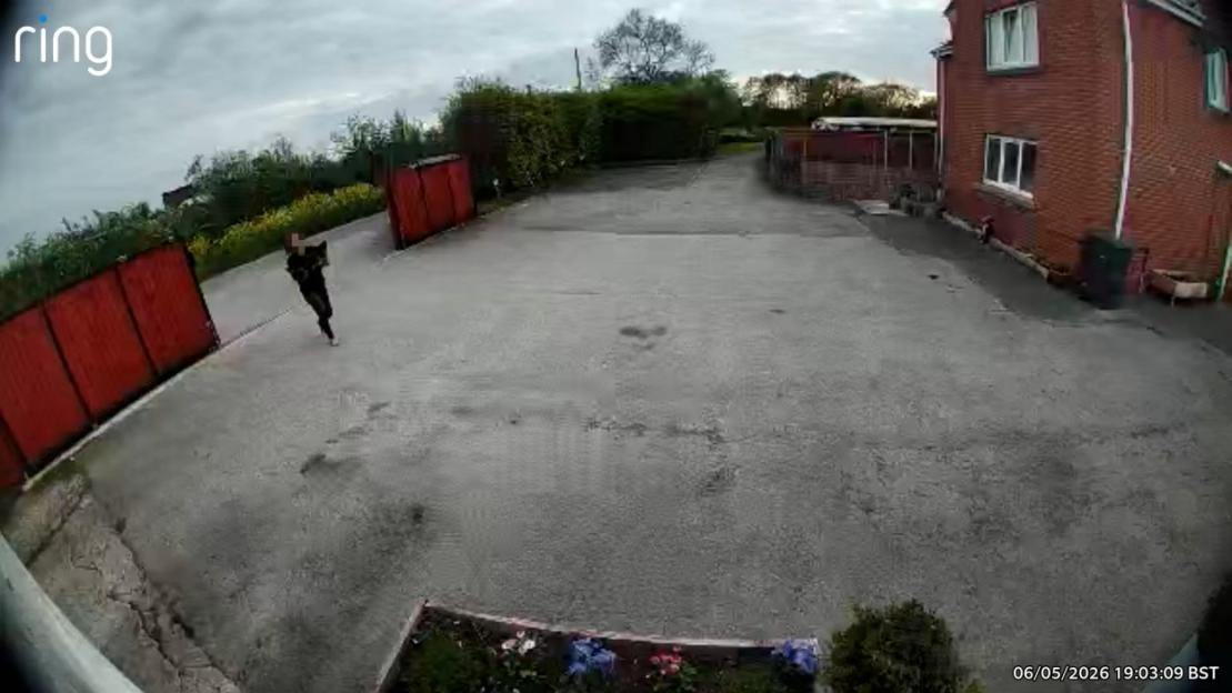 A security camera captures a person running through a driveway, with a red brick house to the right and a red gate on the left.