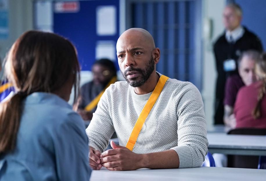 A man with a beard, wearing a light gray sweater and a yellow strap across his chest, sits at a table and talks to a person whose back is to the camera.