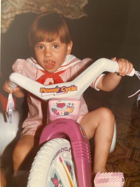 A young child with brown hair, bangs, and a red and white striped top with a red bow, sitting on a pink and white "Power Cycle" tricycle.