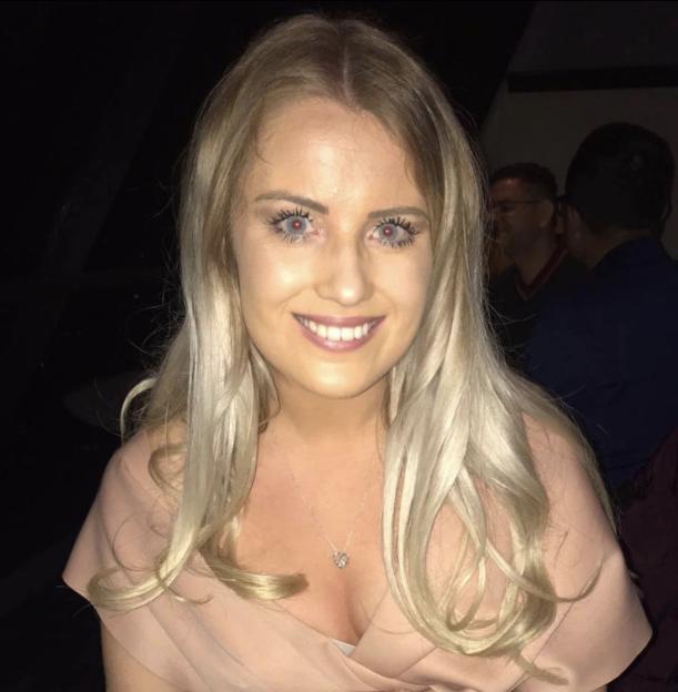 A young woman with long blonde hair and red eyes, possibly from red-eye effect, smiles at the camera.