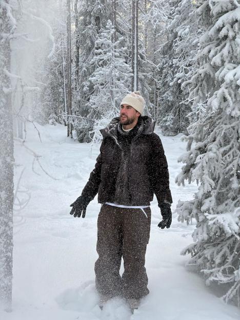 A man standing in a snowy forest, looking upwards as snow falls around him.