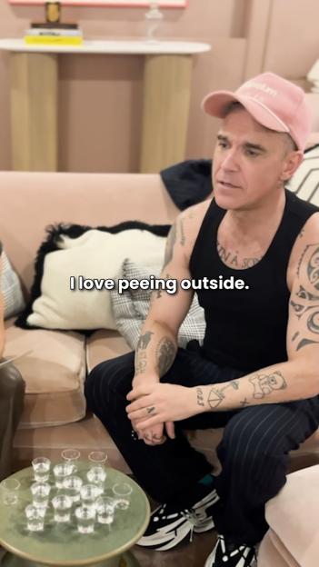 Robbie Williams in a black tank top and pink baseball cap, tattooed arms, sitting on a couch with shot glasses on a table.