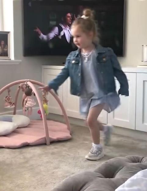 A young girl in a denim jacket dances in front of a TV playing a video.