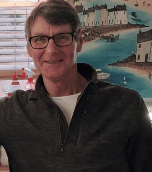 A man smiling in glasses with a painting of a coastal town behind him.