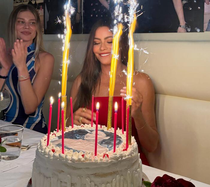Sofia Vergara smiling at her birthday cake with sparklers and candles while a woman claps in the background.