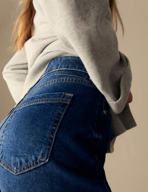 Back view of a person wearing blue jeans with orange stitching and a light grey sweater.