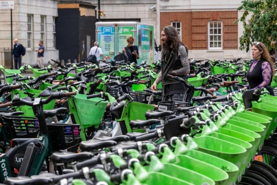 Pedestrians navigate through a dense, disorganized cluster of green and black electric hire bikes parked haphazardly across a public square.