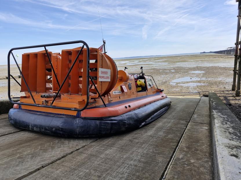 An orange RNLI hovercraft named "Vera Ravine" sits on a concrete ramp leading to a wide, muddy beach at low tide.
