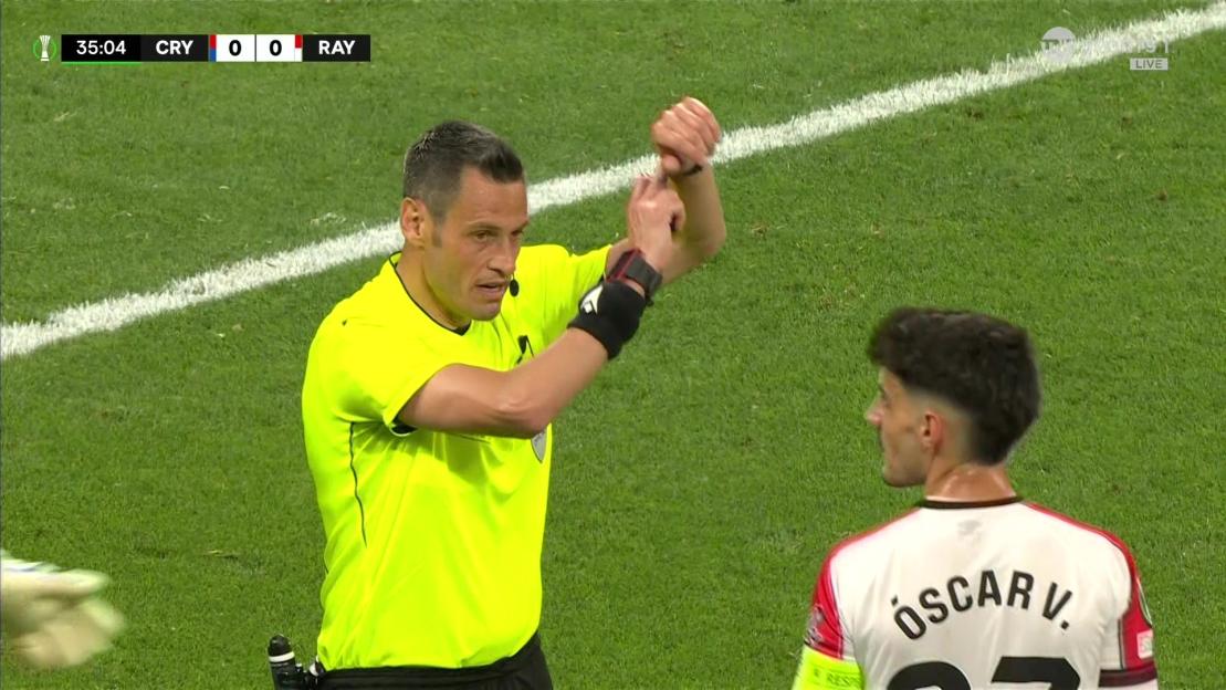 A soccer referee in a neon yellow shirt gestures towards a player.