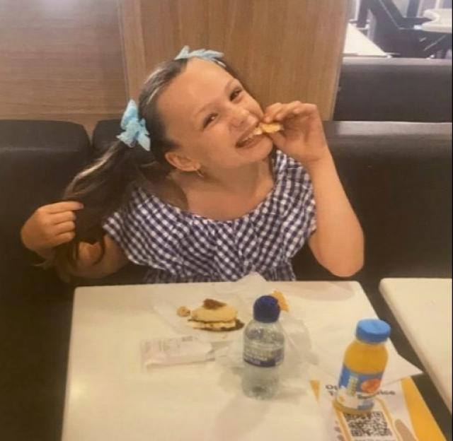 A young girl, Brooke Wiggins, wearing a blue gingham top and blue bows in her pigtails, smiles while eating at a restaurant table.