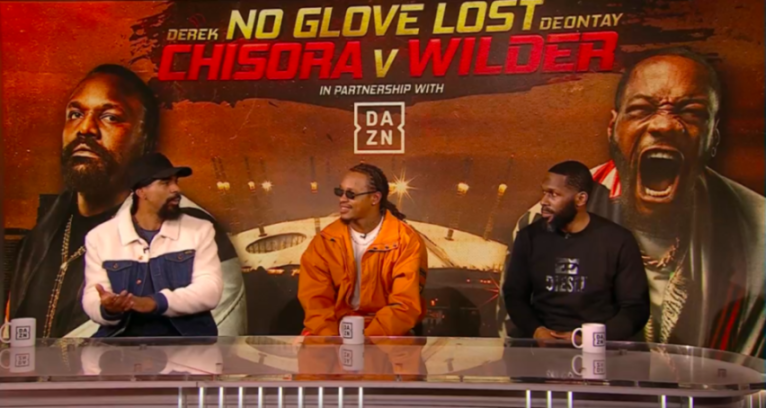Three men sit at a desk in front of a promotional poster for a "No Glove Lost" boxing match featuring Derek Chisora and Deontay Wilder.