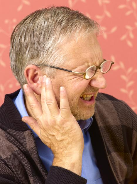 A senior man grimacing with a hand to his ear, suggesting ear pain or tinnitus, against a pink background.