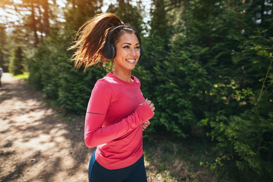 Young woman in athletic wear and headphones jogging on a path lined with trees, smiling.