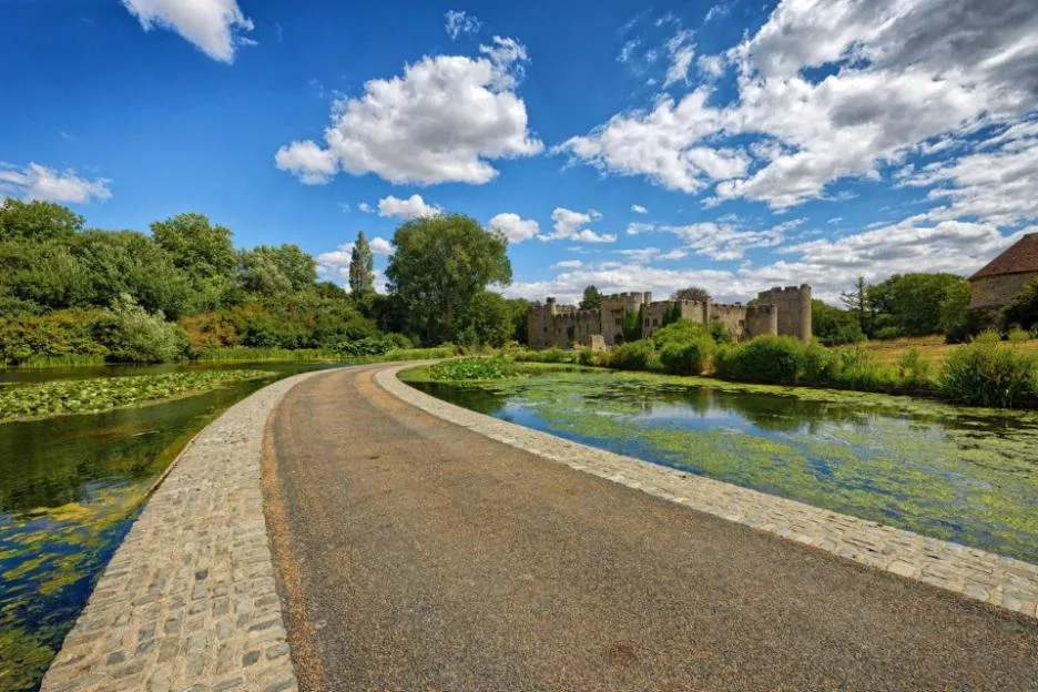 A curved paved path leading across a moat towards a historic stone castle on a sunny day with a blue sky and fluffy white clouds.