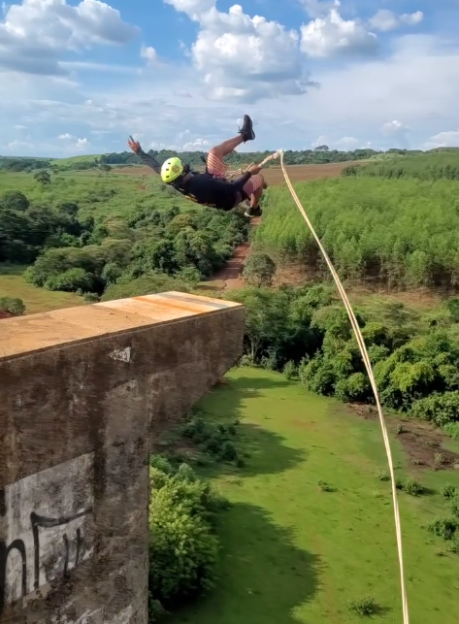 A person bungee jumping off a bridge, suspended in the air with green trees and fields in the background.