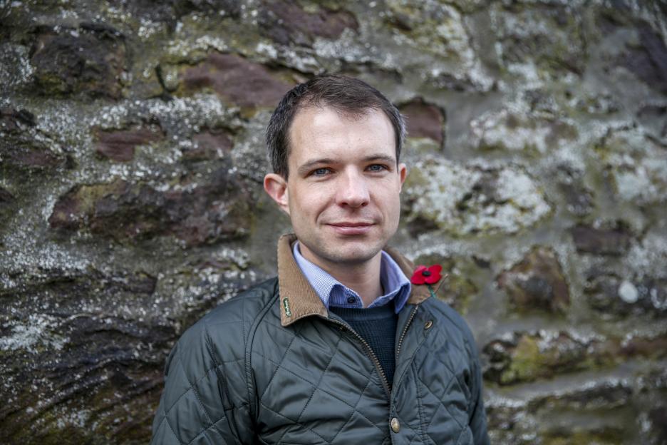 A man in a quilted jacket with a poppy pinned to it, standing against a stone wall.
