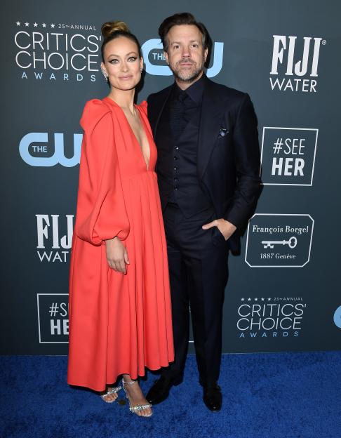 Olivia Wilde in a red dress and Jason Sudeikis in a dark suit pose on the red carpet of the 25th Annual Critics' Choice Awards.