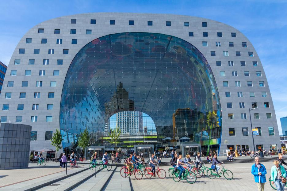 The Market Hall Markthal in Rotterdam, Netherlands, an arched building with a grey stone facade and a glass front, with people cycling and walking outside.