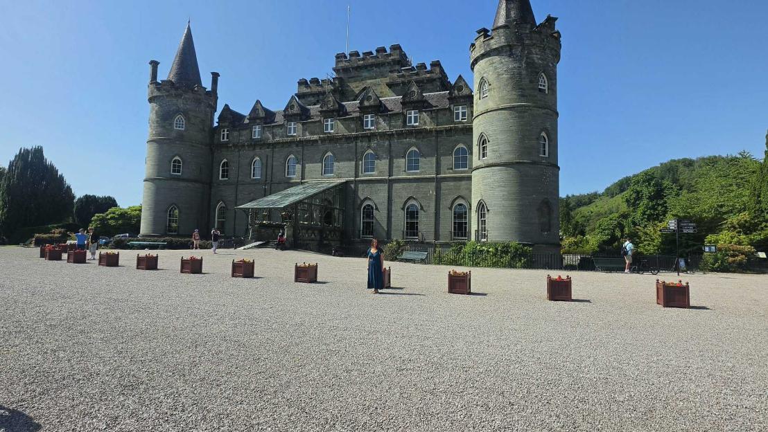 Woman standing in a gravel courtyard in front of a large stone castle with multiple turrets.