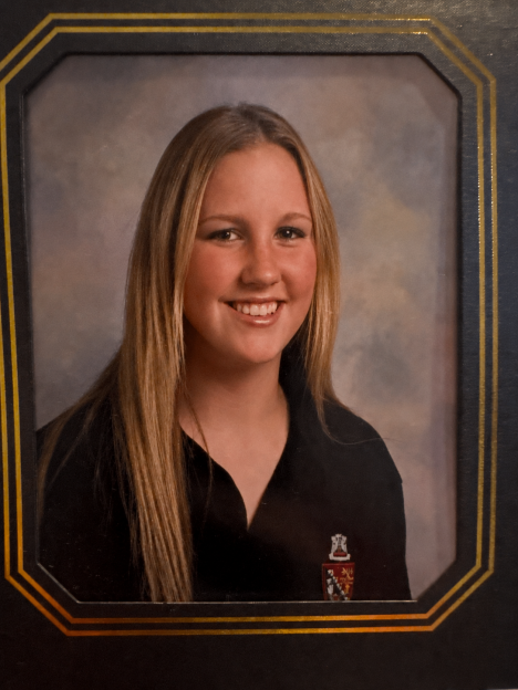 A smiling blonde woman wearing a black polo shirt with a crest on it, framed in a yearbook-style border.