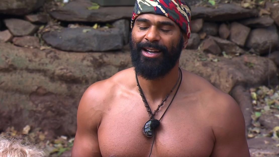 A shirtless man with a beard and a colorful bandana on his head, wearing a necklace with a large pendant.