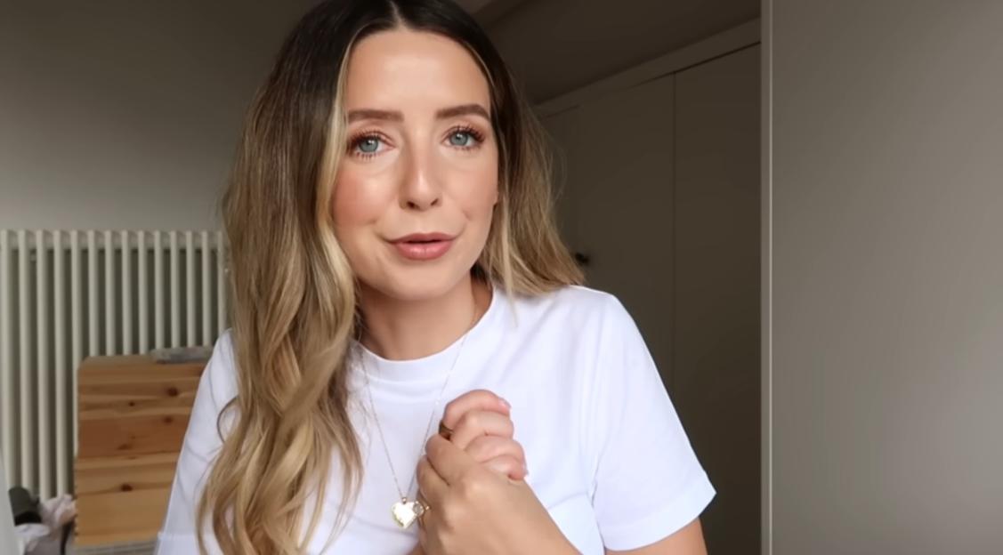 A light-skinned woman with long, wavy blonde hair and blue eyes, wearing a white t-shirt and a heart-shaped locket, looks forward with her hands clasped.