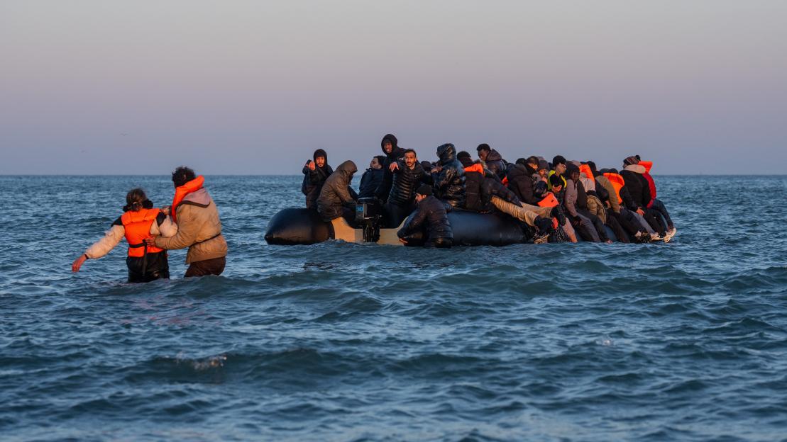 People in an overcrowded dinghy on the sea with two people wading through the water beside the boat.