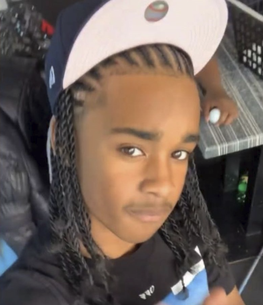 A person with cornrows and long braids wearing a baseball cap looks at the camera.